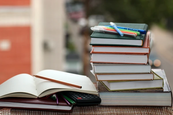 A stack of books and an open notebook with a calculator and pens sit on an outdoor surface with a blurred background.